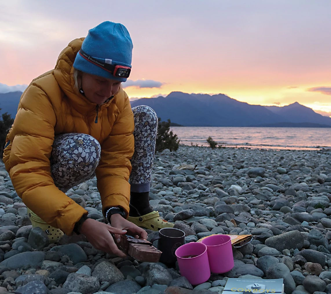 Adventure Guide: Into the fiords, peaks and coastlines of New Zealand’s South Island 3 A camper in a yellow puffer jacket dips a Tim Tam at sunset on a rocky beach